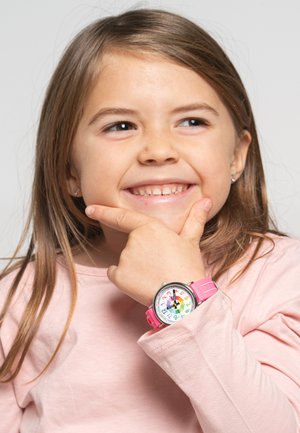 Montre rose avec un cadran coloré comportant des chiffres, portée au poignet d'un enfant. La main de l'enfant repose sur son menton, vêtu d'un t-shirt à manches longues rose clair.