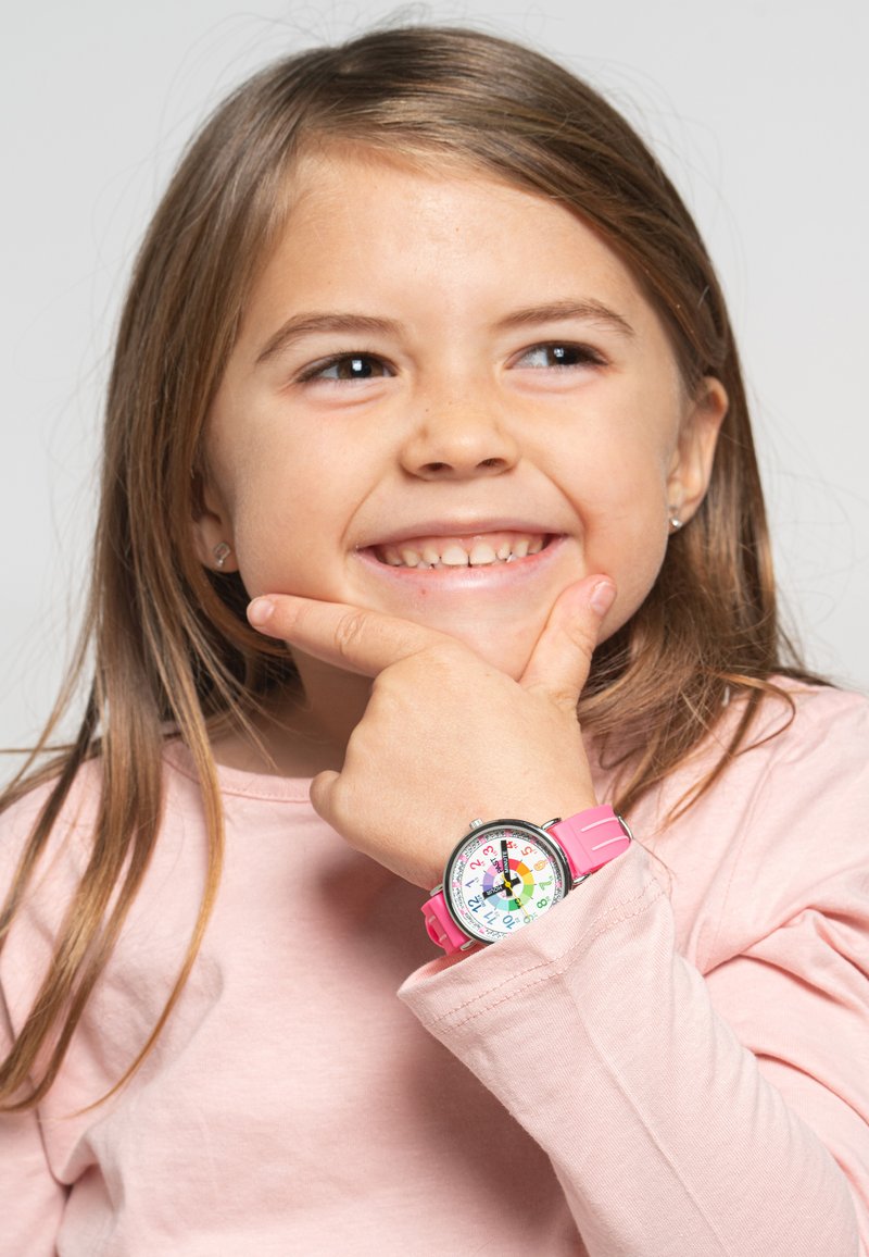 Montre rose avec un cadran coloré comportant des chiffres, portée au poignet d'un enfant. La main de l'enfant repose sur son menton, vêtu d'un t-shirt à manches longues rose clair.
