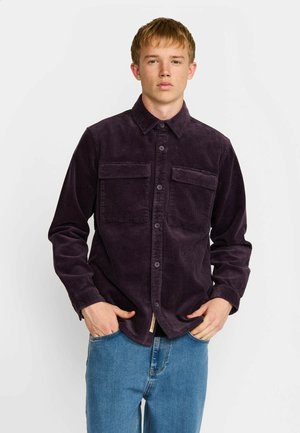 Jeune homme portant une chemise boutonnée en velours côtelé violet foncé avec deux poches poitrine et un jean bleu, debout devant un fond blanc uni.