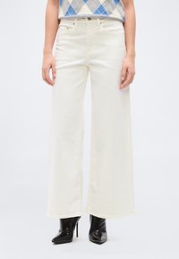 Γυναίκα με κρεμ παντελόνι ψηλόμεσο και φαρδύ στο πόδι, συνδυασμένο με μαύρες μύτες στιλέτο, σε μονόχρωμο φόντο.