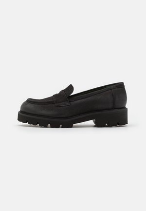 Mocassins en cuir noir avec une surface texturée, un bout arrondi, et une semelle extérieure en caoutchouc. Ils présentent un détail subtil de bride avant et un petit talon.