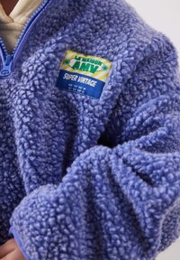 Gros plan d'une veste en polaire bleue avec une fermeture éclair et un patch coloré « LA MAISON AMV SUPER VINTAGE » sur la poitrine.