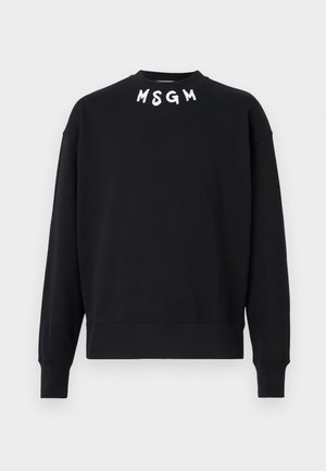 Henkilö, jolla on yllään musta MSGM:n pyöreäpääntiepaita, väljät siniset farkut, mustat kengät ja vasemmalla puolella pieni musta olkalaukku.