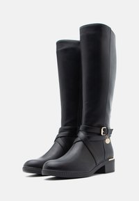 Mustat nahkaiset polven ylle ulottuvat saappaat, joissa on pyöreä kärki, sivuvetoketju ja koristeellinen hihna, jossa on kultainen yksityiskohta. Texturoitu sivupinta. Paksu korko.