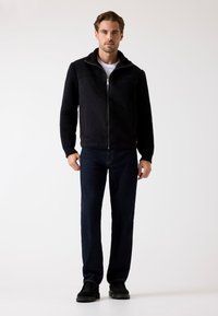 Schwarze Weste mit Reißverschluss, pulloverartige, gerippte Ärmel und ein Kragen aus Lammfell, kombiniert mit dunkelblauen Jeans und schwarzen Schuhen. Glatte Textur.