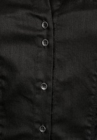 Nærbillede af sort stof med fire lodret placerede sorte knapper, synlig syning langs knaphuller og stofkanter.