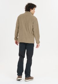 Beige fleece-takk med høy krage, båret over mørke bukser, har et enkelt design. Modellen vises fra ryggen.