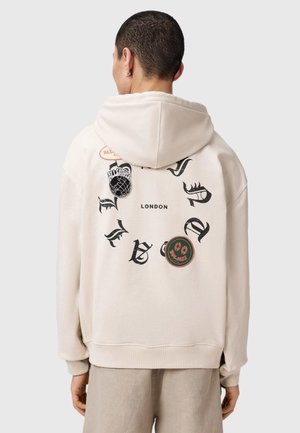 Persoon draagt een beige hoodie met capuchon op, zwarte gotische tekst en badges, waaronder een wereldbol, een smiley en het woord "LONDON" op de achterkant.