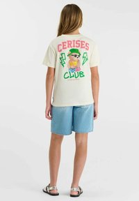 Enfant portant un T-shirt crème avec le texte "Cerises Club" et une figure de dessin animé, associé à un short en denim bleu clair et des sandales argentées.