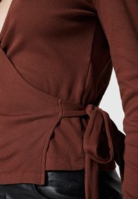 Primer plano de una blusa envuelta de punto con relieve marrón atada en la cintura sobre pantalones de cuero negros de una persona.