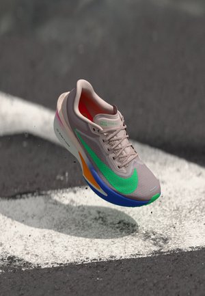 Lys beige løbesko med grøn Nike-swoosh, orange og blå såldetaljer, hængende over sort-hvid tekstureret underlag.