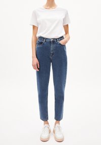 Jean bleu taille haute avec une jambe fuselée, cinq poches et une fermeture à bouton, associé à un t-shirt en coton blanc uni et des baskets blanches.