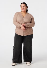 Lichtbruin cardigan met een V-hals en gouden knoopsluiting, gecombineerd met wijde zwarte spijkerbroek en witte instapschoenen met studs.