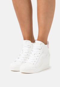 Sneakers bianche alte con suola in gomma strutturata, chiusura frontale con lacci e marchio discreto sul tallone. Finitura liscia con un design elegante.