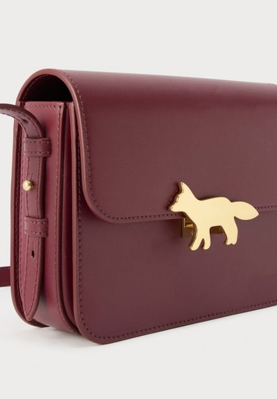 Σκούρα κόκκινη δερμάτινη τσάντα χειρός με ρυθμιζόμενο λουράκι και χρυσό κούμπωμα σε σχήμα αλεπούς στο εμπρός καπάκι, εμφανιζόμενη σε λευκό φόντο.