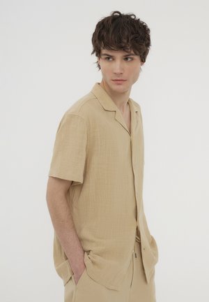 Camicia beige a maniche corte con bottoni, realizzata in tessuto strutturato, dotata di colletto e vestibilità ampia, abbinata a pantaloni coordinati.