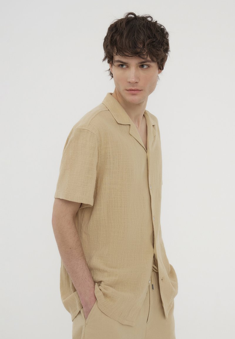 Camicia beige a maniche corte con bottoni, realizzata in tessuto strutturato, dotata di colletto e vestibilità ampia, abbinata a pantaloni coordinati.