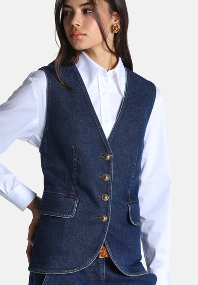 Femme portant un gilet en jean bleu foncé sur une chemise blanche à boutons, avec des boucles d'oreilles dorées et les mains posées sur les hanches.