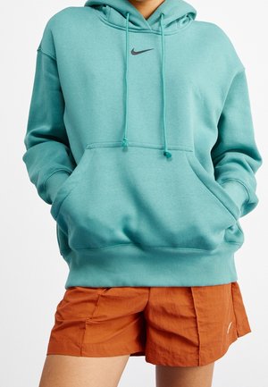 Osoba nosząca turkusową bluzę z kapturem Nike z przednią kieszenią i spalonymi pomarańczowymi szortami, dłonie częściowo w kieszeniach bluzy, na białym tle.