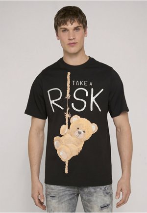 Noor mees kannab musta T-särki, millel on kaisukaru ronimas köit mööda ja ees tekst "Take a Risk".