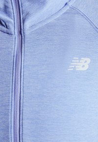 Ljusblå zip-up hoodie i texturerat tyg med en liten vit New Balance-logotyp på vänster bröstområde.