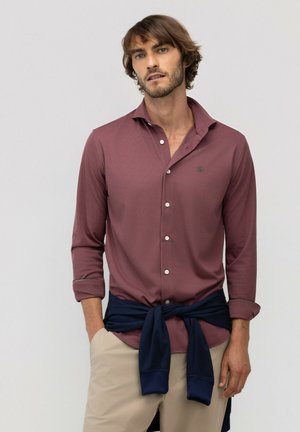 Hombre con cabello castaño despeinado lleva una camisa burdeos abotonada, pantalones beige y un suéter azul marino atado alrededor de su cintura con un fondo liso.
