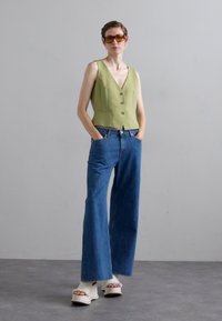 Top sin mangas de color verde claro con botones al frente, combinado con jeans azul de tiro alto. La modelo lleva sandalias de plataforma blancas y gafas de sol.