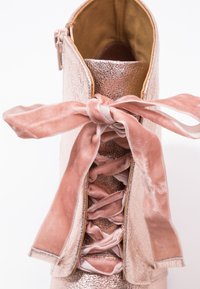 Stivaletto rosa oro con finitura metallica, caratterizzato da una parte superiore piegata, lacci in velluto e una morbida fodera interna. I lacci sono legati a fiocco.