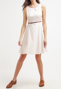 Un vestido sin mangas de lunares en beige claro, con un cuerpo ajustado, una falda A-line suave y un cinturón marrón en la cintura.
