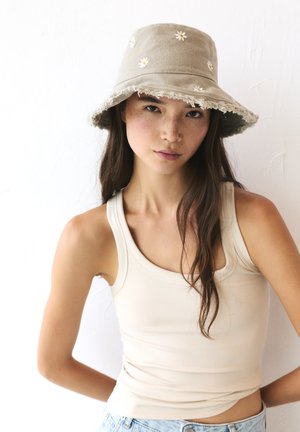 Cappello a secchiello color khaki con bordi sfrangiati, decorato con ricami di margherite bianche. Il modello indossa una canotta beige, con texture e colori naturali e morbidi.