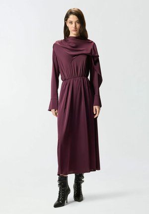 Haljina duboko ljubičaste boje s dugim rukavima, skupljena u struku, s naboranim izrezom i lepršavim krojem, uparena s crnim gležnjačama na vezanje od čipke.