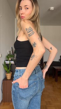 Jeune femme aux cheveux blonds portant un débardeur noir et un jean bleu, montrant des tatouages sur ses bras, debout dans un salon.