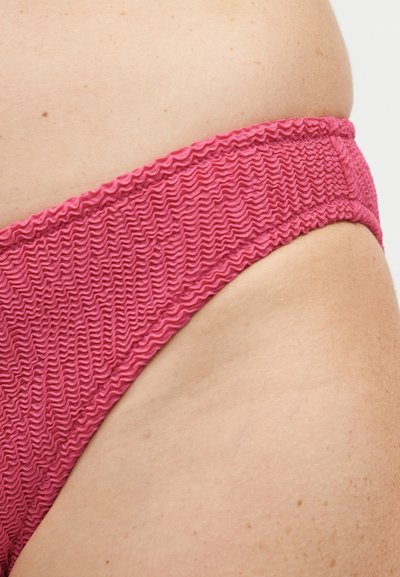 Κοντινό πλάνο του ισχίου ενός ατόμου που φοράει ροζ μαγιό με υφή και κυματιστό σχέδιο, σε φωτεινό φόντο.