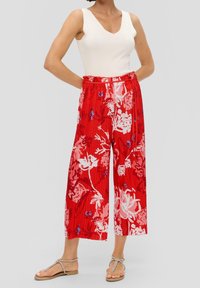 Femme portant un haut sans manches blanc, un pantalon large cropped rouge à fleurs, et des sandales plates beiges à lanières, debout les mains dans les poches.