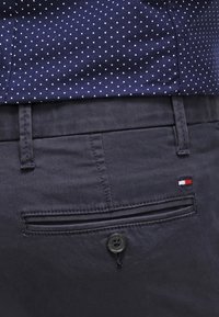 Pantalons bleu marine foncé avec une poche arrière dotée d'une fermeture à bouton. Le tissu semble lisse avec une texture subtile ; détail du logo en rouge, blanc et bleu.