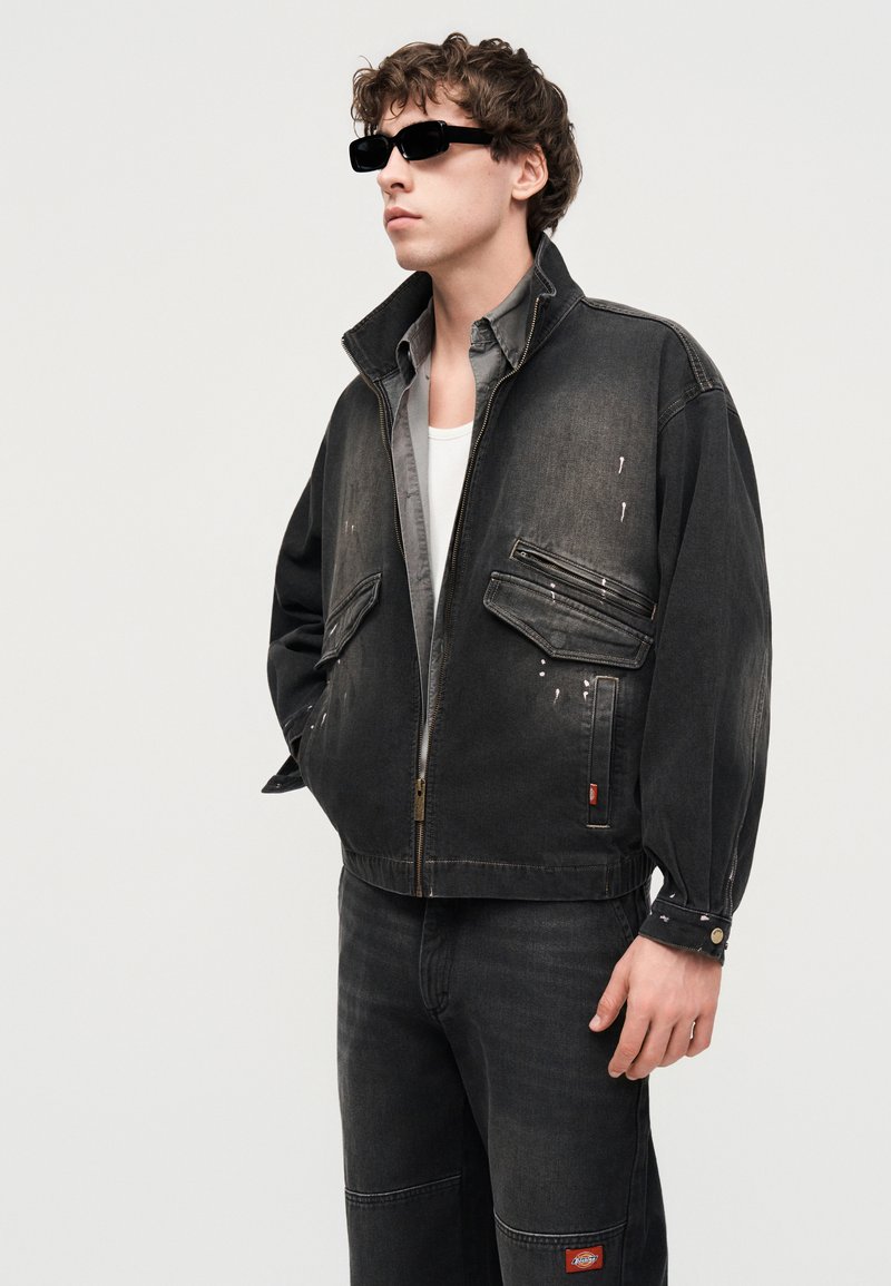 Giovane uomo che indossa una giacca nera sbiadita oversize con tasche frontali, jeans neri, camicia bianca e occhiali da sole rettangolari neri, in piedi di lato.