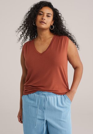 Kvinde med krøllet hår iført rustfarvet, ærmeløs top og lyseblå bukser med snøre, poserer med den ene hånd i lommen.