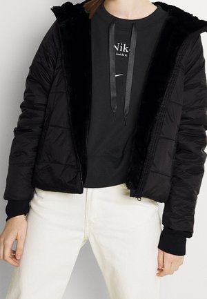 Personne portant une doudoune noire doublée de fourrure par-dessus un sweat Nike noir et un pantalon blanc, coupée à mi-torse.