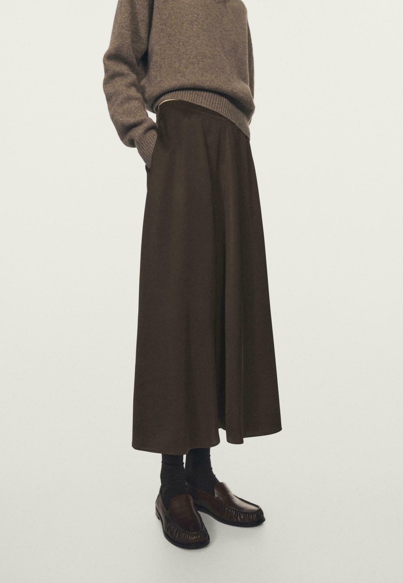 Personne portant un pull marron et une longue jupe marron foncé avec des chaussettes noires et des mocassins marron, debout devant un fond clair uni.