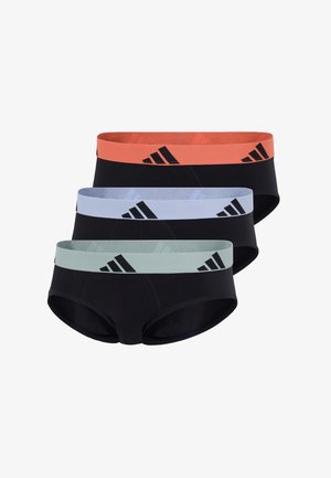 Athletischer Mann steht barfuß und trägt schwarze Slips mit einem orangen Taillenbund, der drei schwarze Adidas-Logos zeigt, vor weißem Hintergrund.