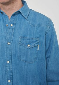 Camicia di denim di colore azzurro chiaro, con colletto a punta, chiusura anteriore con bottoni madreperlacei e taschino sul lato sinistro del petto.