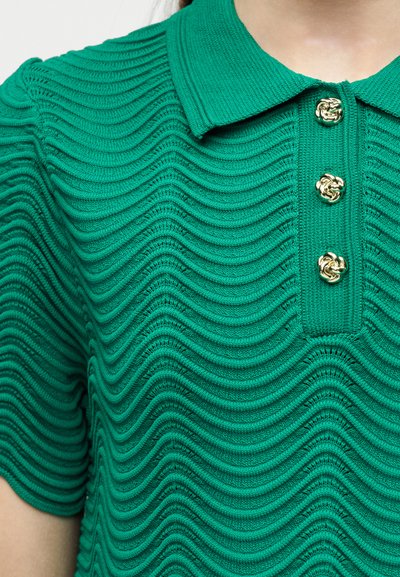 Πράσινο πλεκτό πόλο πουκάμισο με υφασματώδη κυματιστά μοτίβα και τρία διακοσμητικά χρυσά κουμπιά στην πατιλέτα, φορεμένο από ένα άτομο.