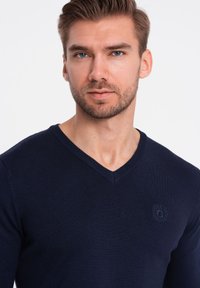 Mörkblå v-ringad tröja tillverkad av ett strukturerat stickat tyg, med en diskret emblem på bröstet och åtsittande ärmar.