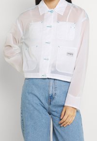 Giacca trasparente bianca corta con accenti blu, caratterizzata da una chiusura con bottoni, grandi tasche e un colletto. Indossata con jeans azzurri.