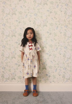 Jeune fille aux longs cheveux noirs portant une robe crème avec des nœuds rouges, des chaussettes bleues et des chaussures marron, debout devant un papier peint à motif floral.