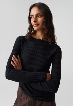 Femme aux longs cheveux bruns, portant un haut noir à manches longues côtelé et un pantalon marron, les bras croisés, regardant sur le côté devant un fond gris.