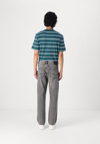 Camisa de manga corta a rayas en tonos verde azulado y blanco combinada con pantalones de mezclilla grises; costuras visibles y bolsillos traseros, llevada con zapatillas blancas.