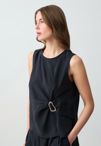 Top senza maniche nero con design frontale attorcigliato, dotato di una chiusura decorativa in metallo e tessuto liscio e leggero.