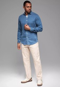 Jeanshemd in Mittelblau mit klassischem Kragen und langen Ärmeln. Kombiniert mit hellbeigen Hosen und braunen sowie weißen Sneakers.