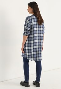 Langärmliges karierter Hemd in Marineblau, Weiß und Grün. Mit hochgerollten Ärmeln, einem abgerundeten Saum und einer lockeren Passform, kombiniert mit dunklen Jeans und schwarzen Schuhen.